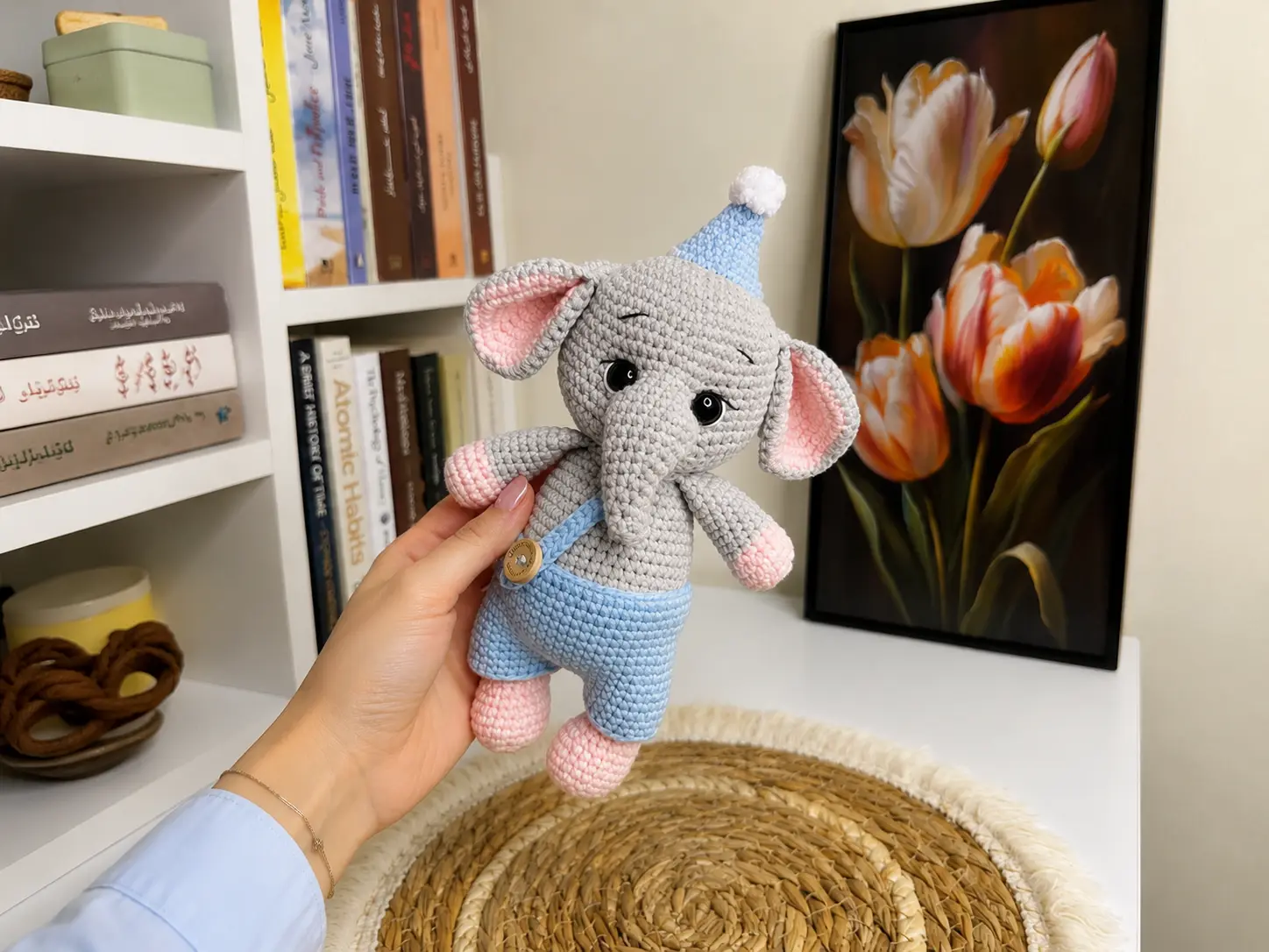 Hand holding grey crochet baby elephant amigurumi in blue overalls with books and tulip art in background