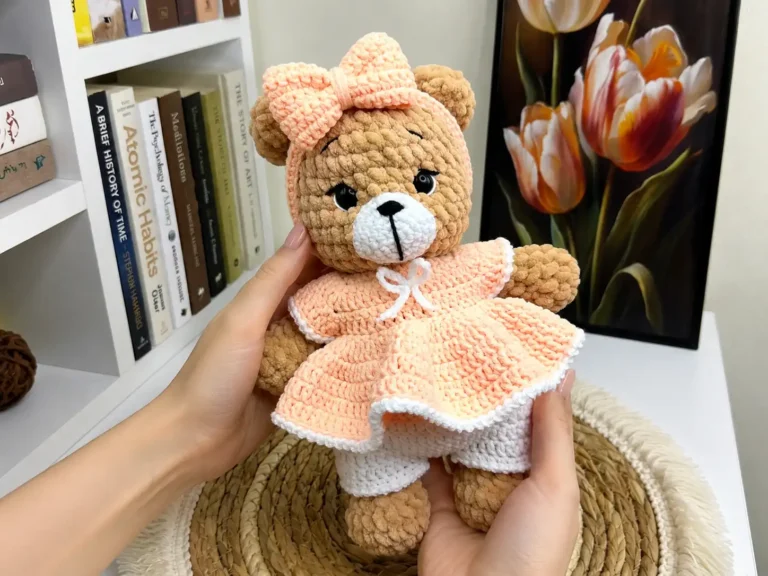 Hands holding a plush brown crochet bear in a peach dress and bow, featured in a crochet bear amigurumi free pattern.