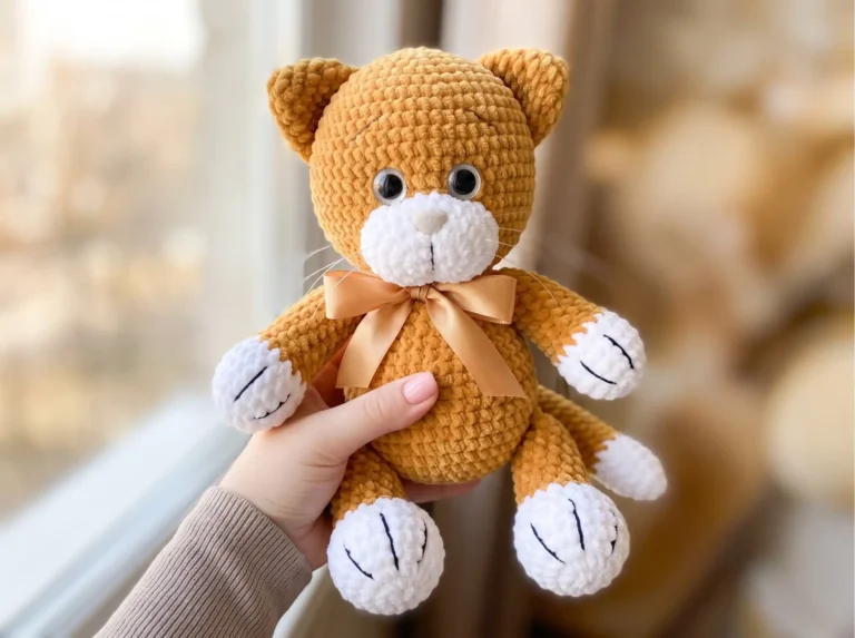 Ginger chunky yarn crochet cat amigurumi free pattern toy with white paws and a ribbon, held by a bright window.