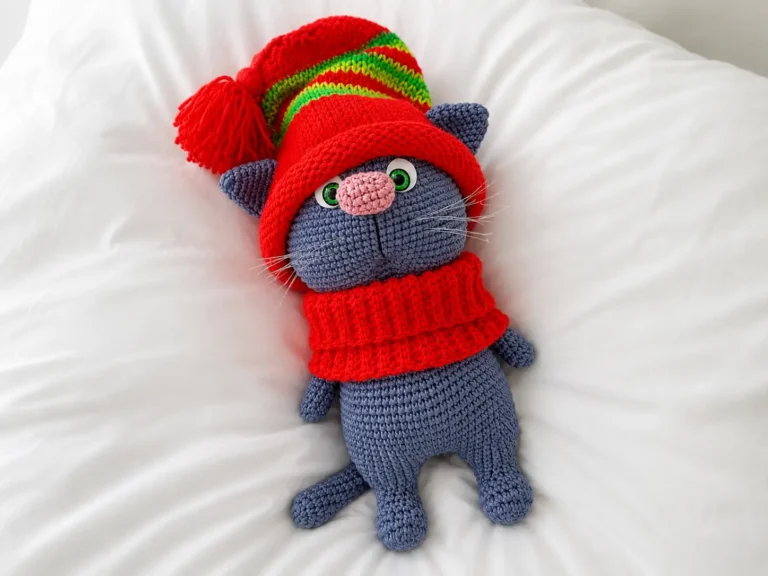 A grey toy made from our crochet cat amigurumi free pattern, wearing a red hat and scarf, lying on a white sheet.