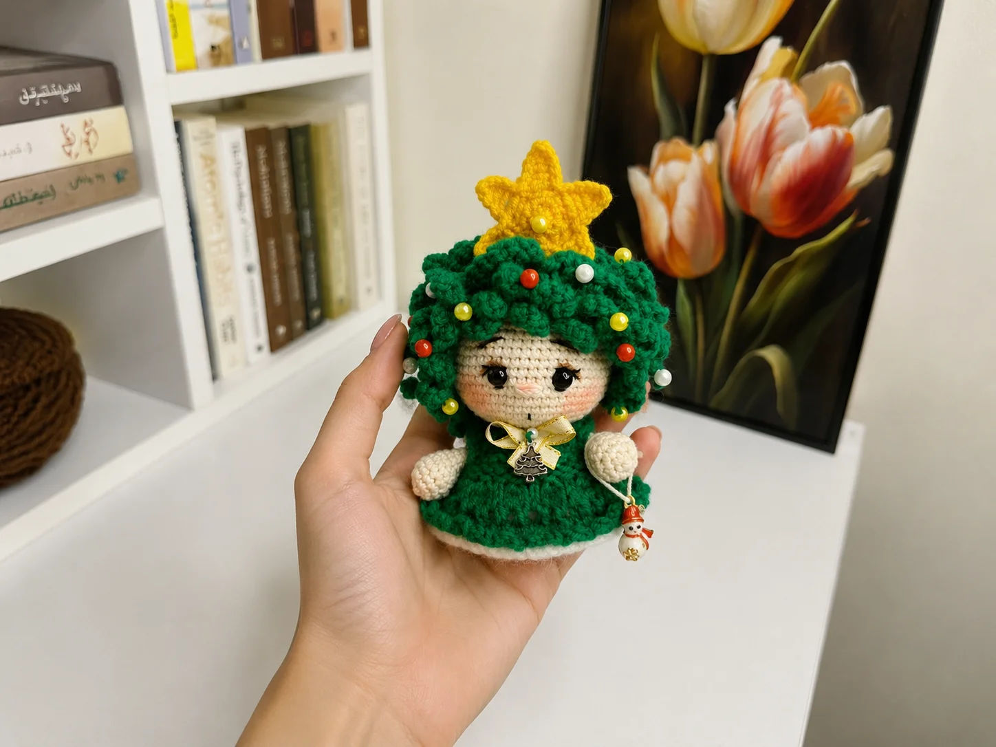 Hand holding a green crochet christmas tree amigurumi doll with yellow star and colorful beads against bookshelf background