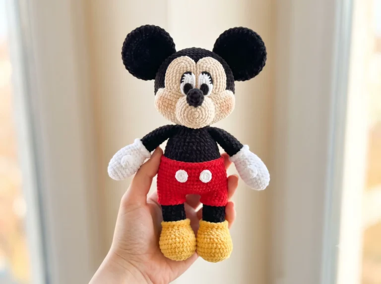 Hand holding a chunky yarn crochet classic cartoon mouse pattern toy in black, red, and yellow against a blurred background.