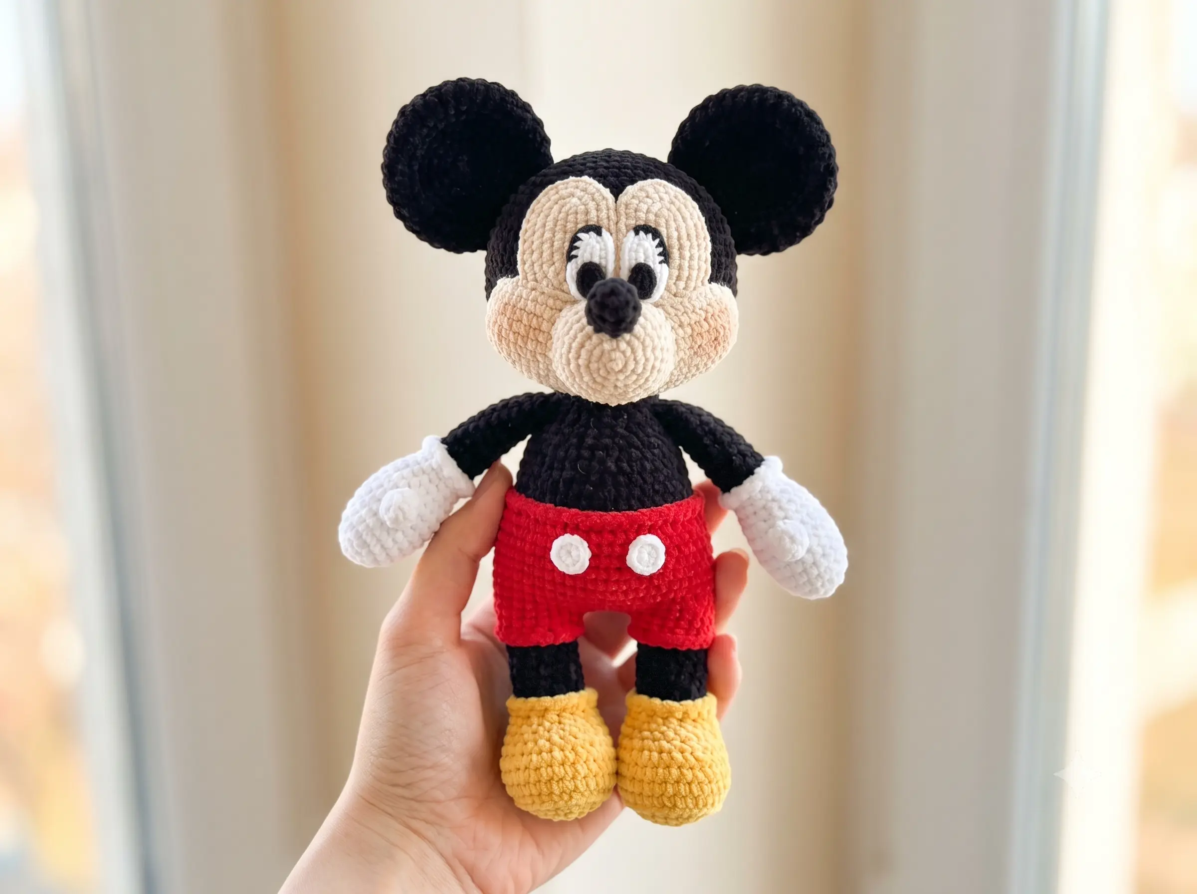 Hand holding a chunky yarn crochet classic cartoon mouse pattern toy in black, red, and yellow against a blurred background.