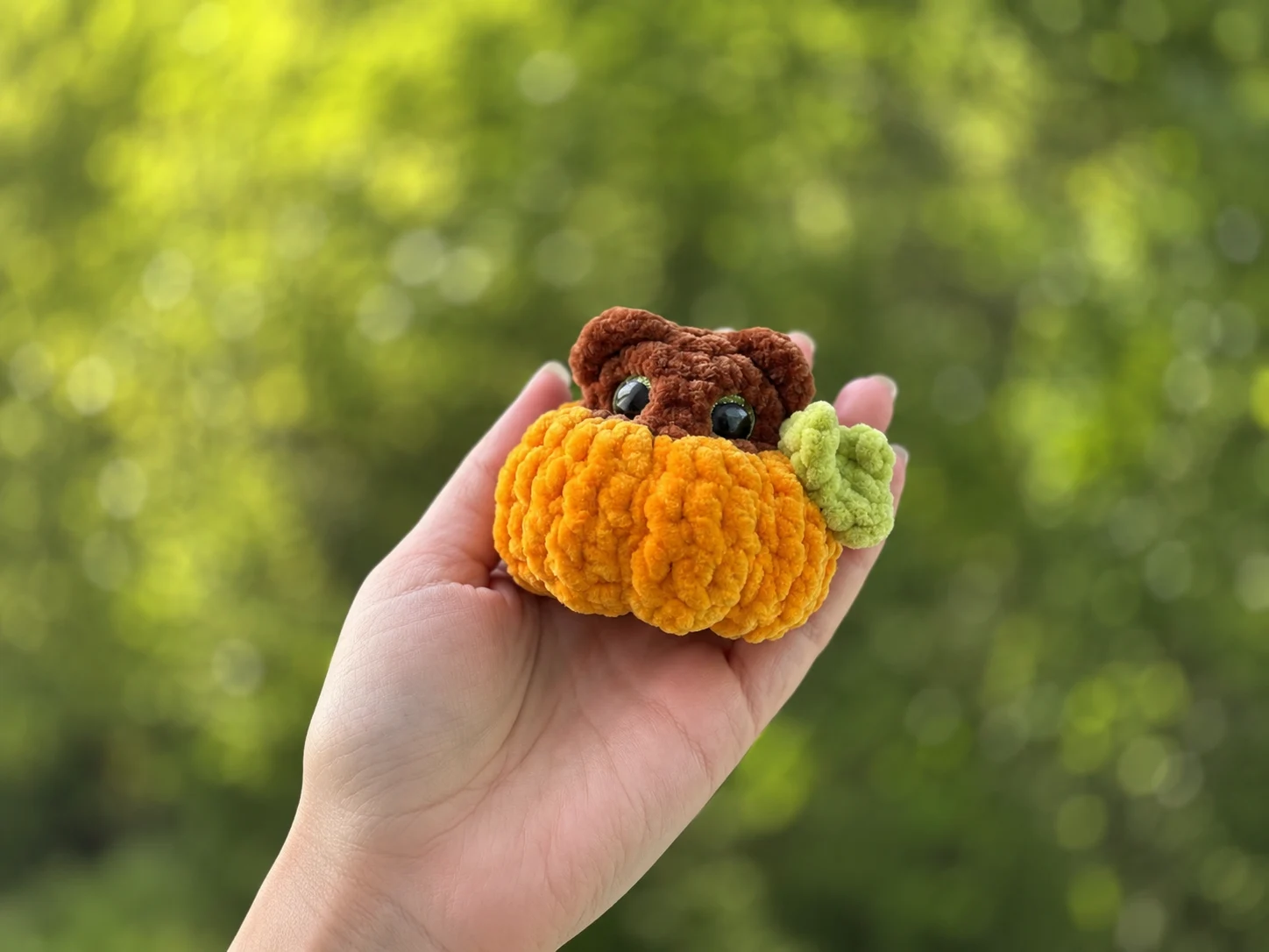 Hand holding a plush orange velvet crochet halloween pumpkin free pattern bear toy outdoors against a blurry green background.
