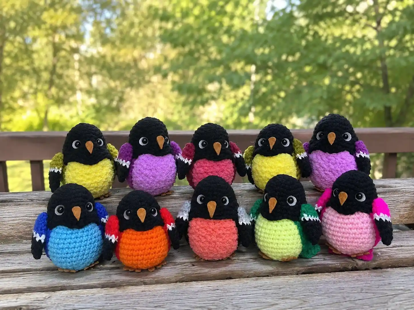 Group of colorful crocheted birds with black heads lined up on a wooden railing outdoors.