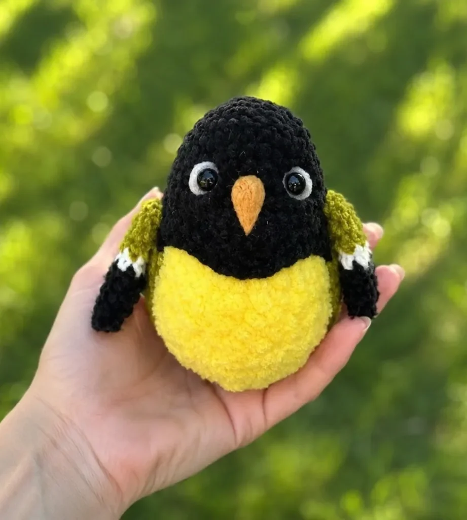 Hand holding a tiny yellow and black crocheted bird outdoors with blurred green background.