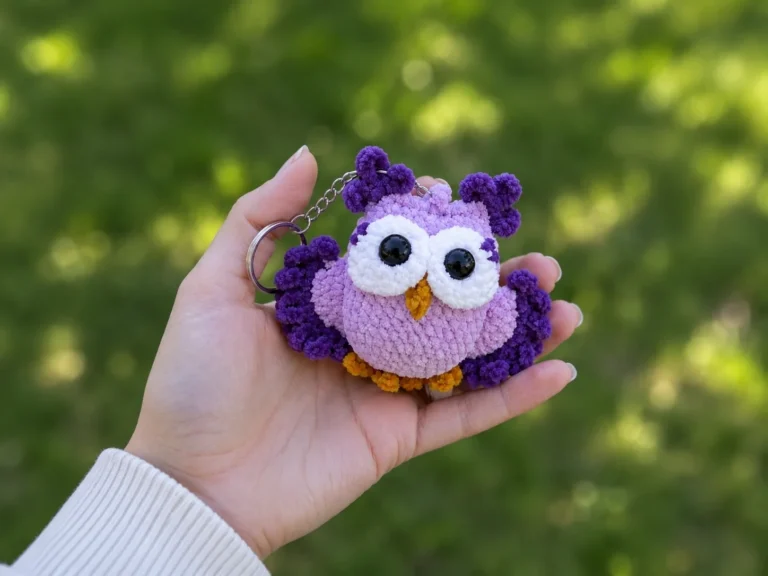 Hand holding a fluffy purple crochet owl keychain free pattern with yellow feet against a blurred green background.