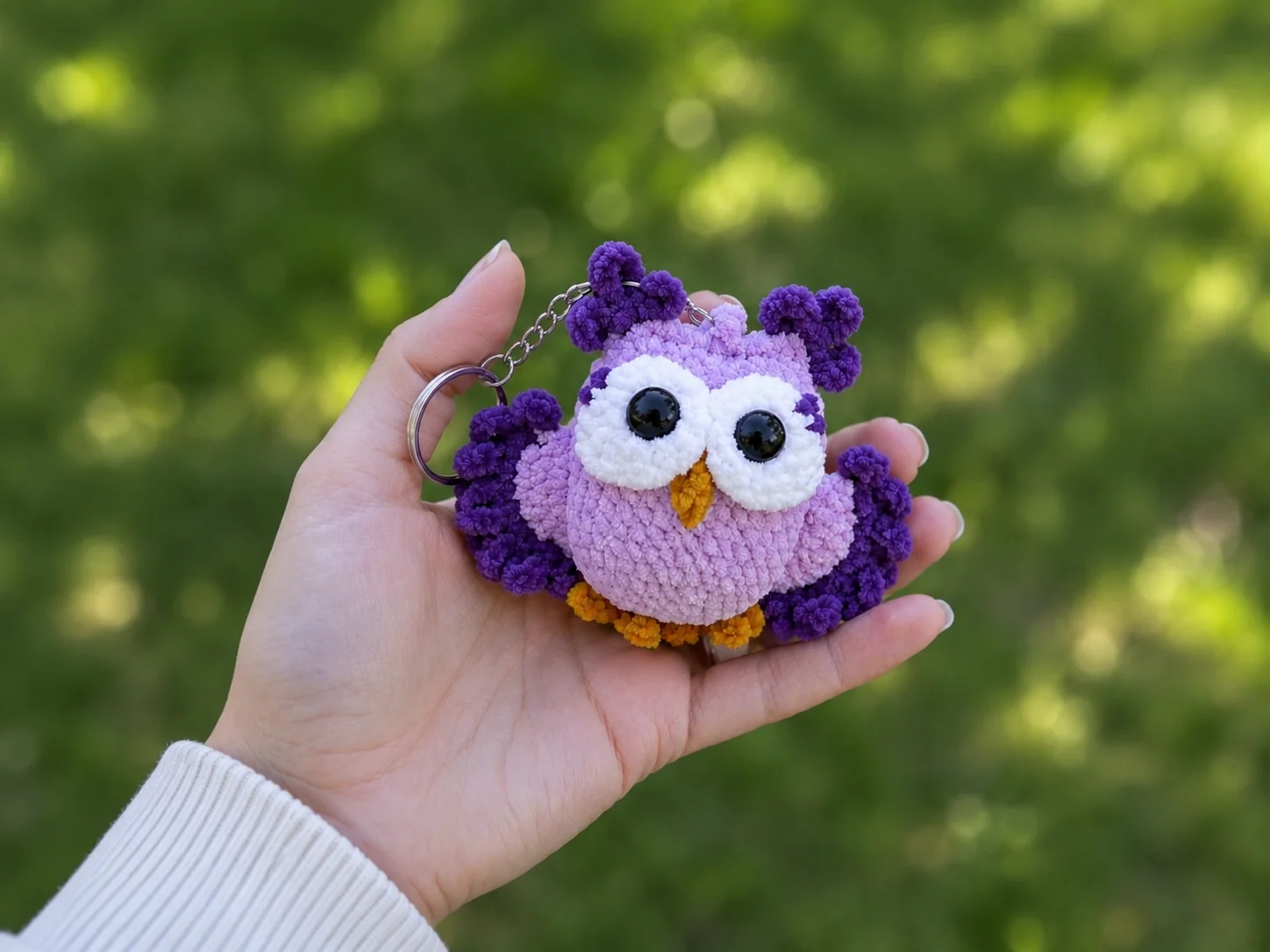 Hand holding a fluffy purple crochet owl keychain free pattern with yellow feet against a blurred green background.