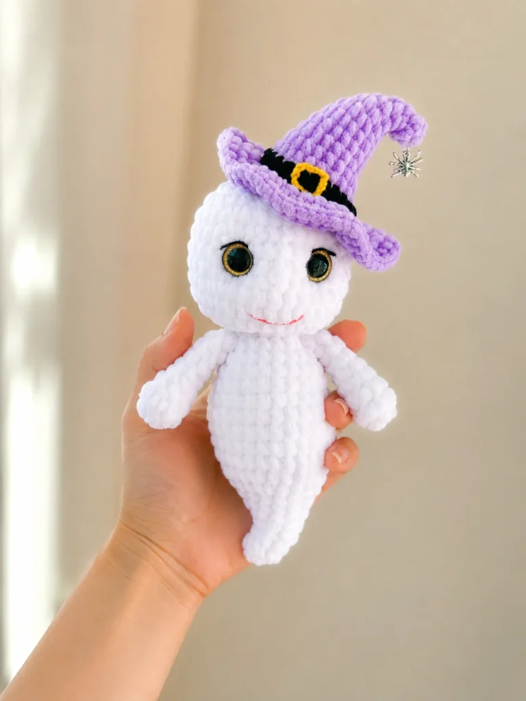 A hand displays a soft white amigurumi ghost wearing a purple witch hat, with a blurry green yard background.