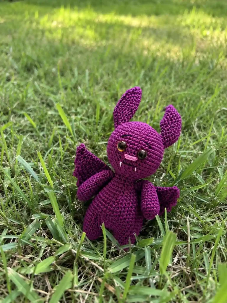 Finished purple crocheted Halloween bat plushie with scalloped wings and tiny fangs standing on green grass.
