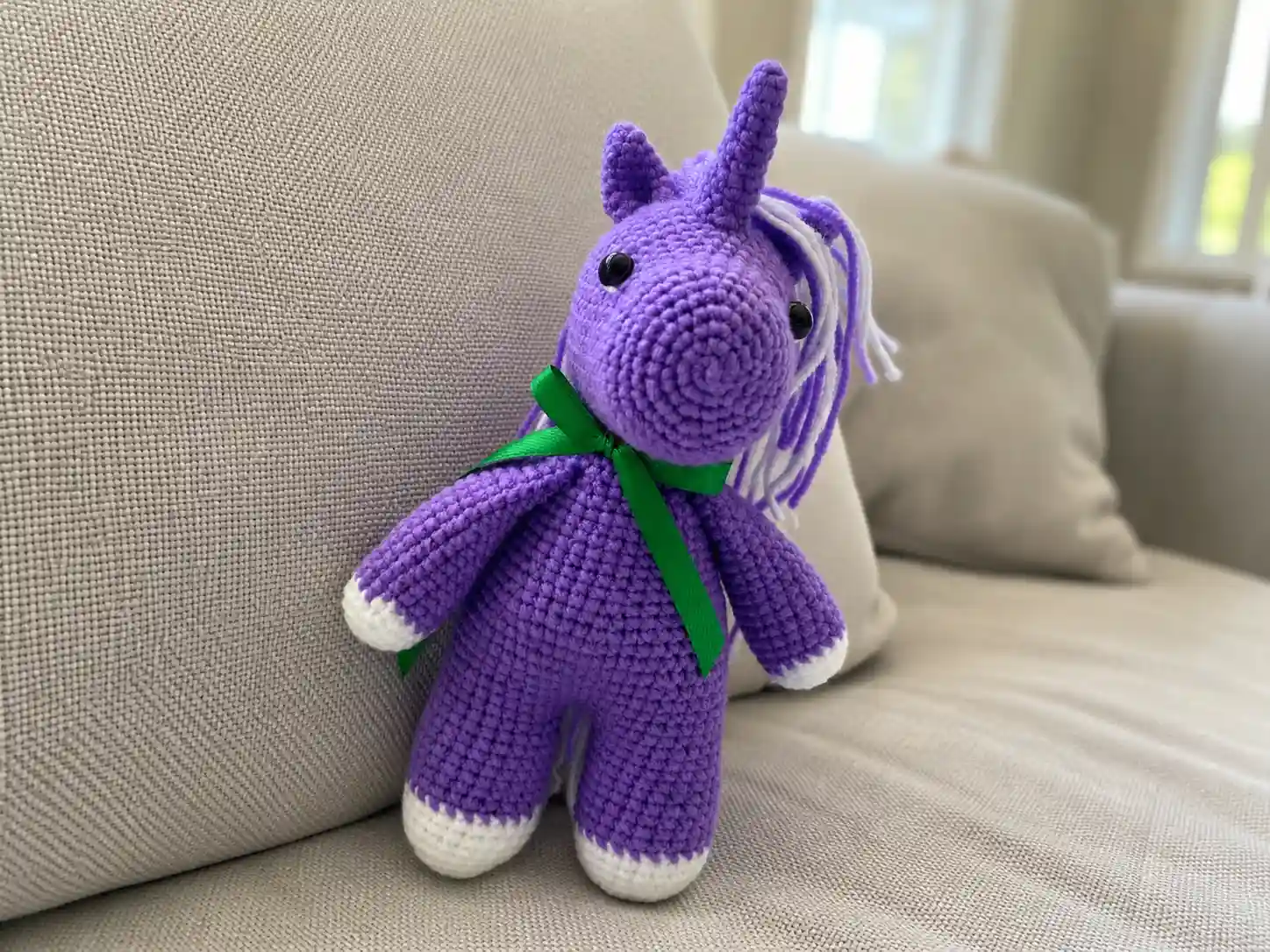 Purple yarn toy from a crochet unicorn amigurumi free pattern with a green ribbon, sitting on a beige couch.