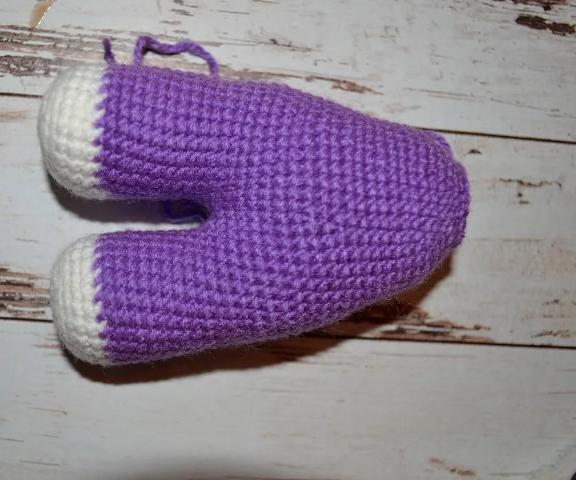 The lower half of a crocheted amigurumi unicorn resting on a wooden table, featuring two violet legs with white hooves joined into a continuously worked, tapered violet body.