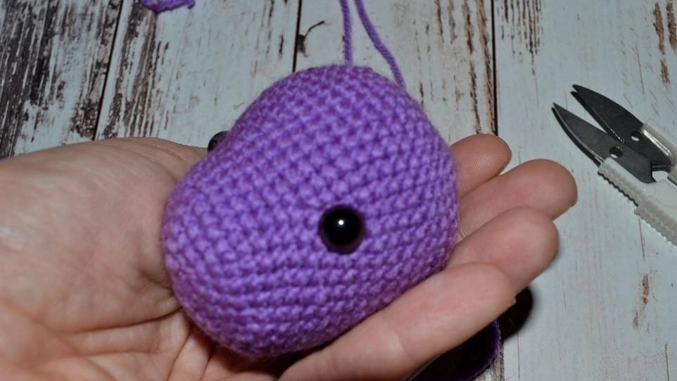 A close-up of the crocheted head of Viola the unicorn being held in a hand, showcasing the placement of the black safety eyes on the violet yarn.