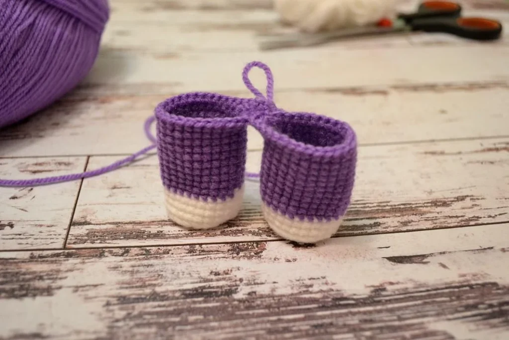 Two violet and white crocheted unicorn legs standing upright on a rustic wooden surface, connected at the top by a small two-stitch yarn chain.