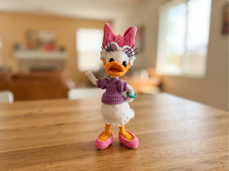 Crochet daisy duck amigurumi with pink bow, purple top, and white skirt on a wooden table in a blurred living room.