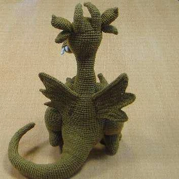 A back view of a completed green crochet dragon amigurumi, displaying the attachment of its angled, feathered wings, the placement of four head appendages (ears and horns), and a thick, wire-posed tail curving to the left.