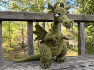 Green winged dragon amigurumi free pattern sitting on a wooden rail against a blurred forest backdrop.