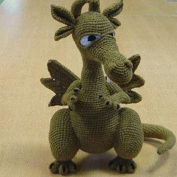 A finished green crochet dragon amigurumi standing upright, featuring detailed wings, horns, white eyes with black pupils, and a poseable wire-framed tail.