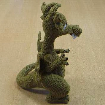 A side profile view of a finished green crochet dragon standing upright, showcasing its open jaw with white fangs, curved wire-framed tail, thick hind legs, and angled wings.