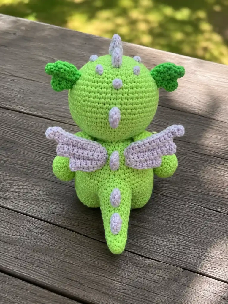 Back view of a finished light green crocheted amigurumi dragon showing lilac wings, back spikes, and tail