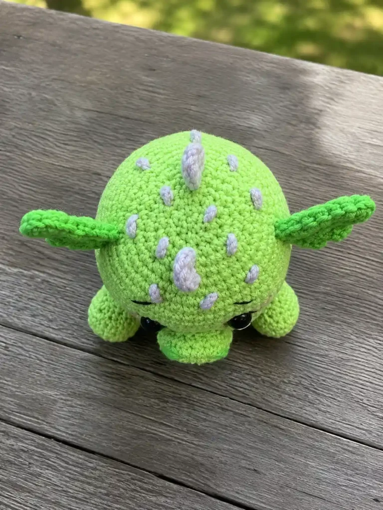 Top-down view of a light green crocheted amigurumi dragon showing head details, purple French knot accents, and back spikes