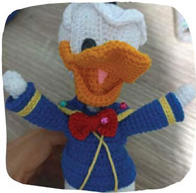 A finished, handmade amigurumi duck with a white head, orange beak, and wearing a blue sailor-style jacket with yellow trim and a red bow tie.