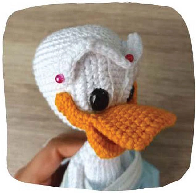 A close-up view of the head of a handmade amigurumi duck, featuring a crisp white crocheted head, a bright orange beak, black safety eyes, and decorative pink accents on the white cap.