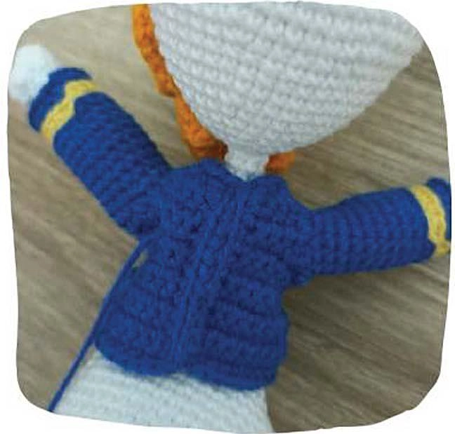 A back view of the amigurumi duck, showcasing the blue crocheted sailor-style jacket with yellow sleeve trim and the poseable arms.