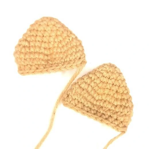 Two flat, triangular tan crocheted amigurumi cat ears with long yarn tails left attached.