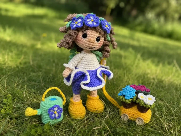 Crochet flower fairy amigurumi doll in blue dress standing on green grass next to a tiny watering can and flower cart.