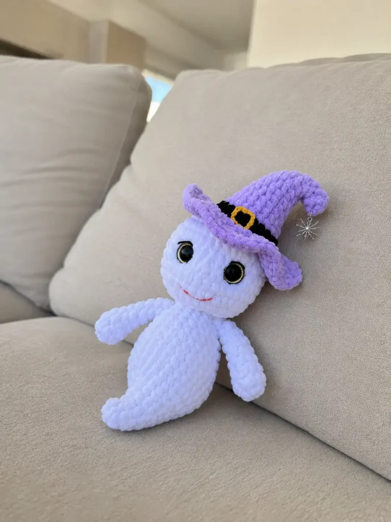 Crocheted white Halloween ghost amigurumi made with plush chenille yarn, wearing a purple witch hat with a yellow buckle.