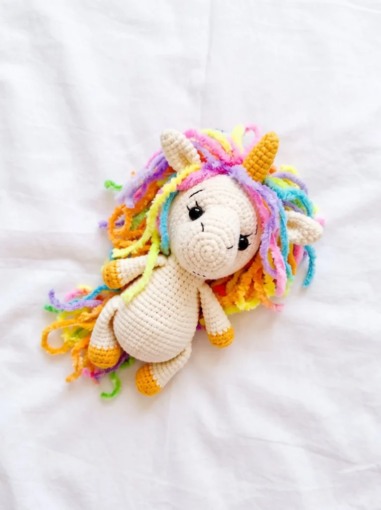 finished cream-colored crochet unicorn lying on a white sheet, representing the free unicorn amigurumi pattern with horn and yarn mane.