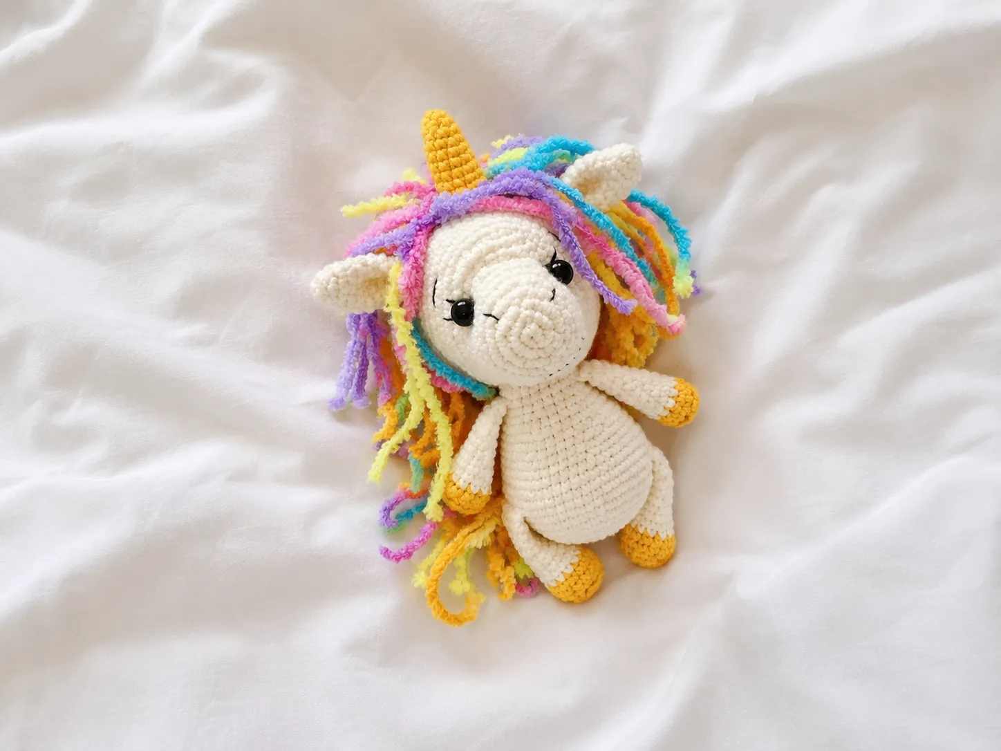 A cream crochet toy with a fuzzy rainbow yarn mane lying on a white bedsheet for this free unicorn amigurumi pattern.