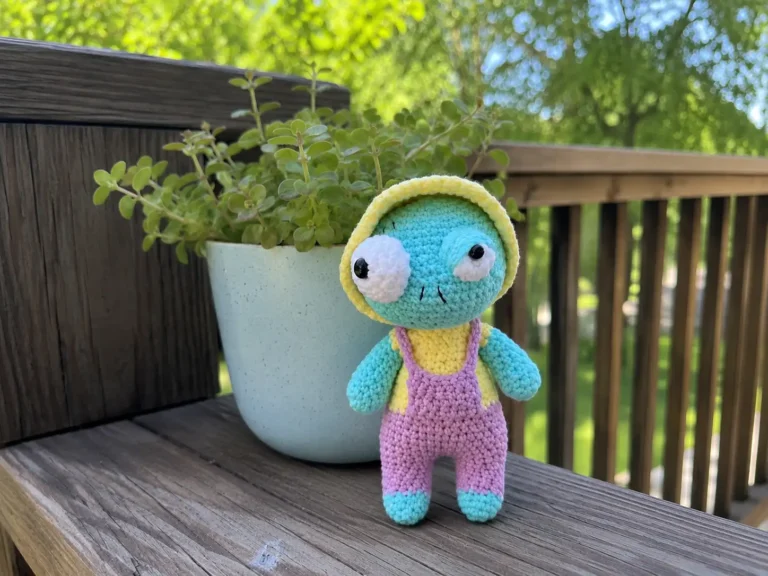 A moss-green crochet zombie doll in a brick-red hooded sweater and charcoal pants, sitting on a shelf.
