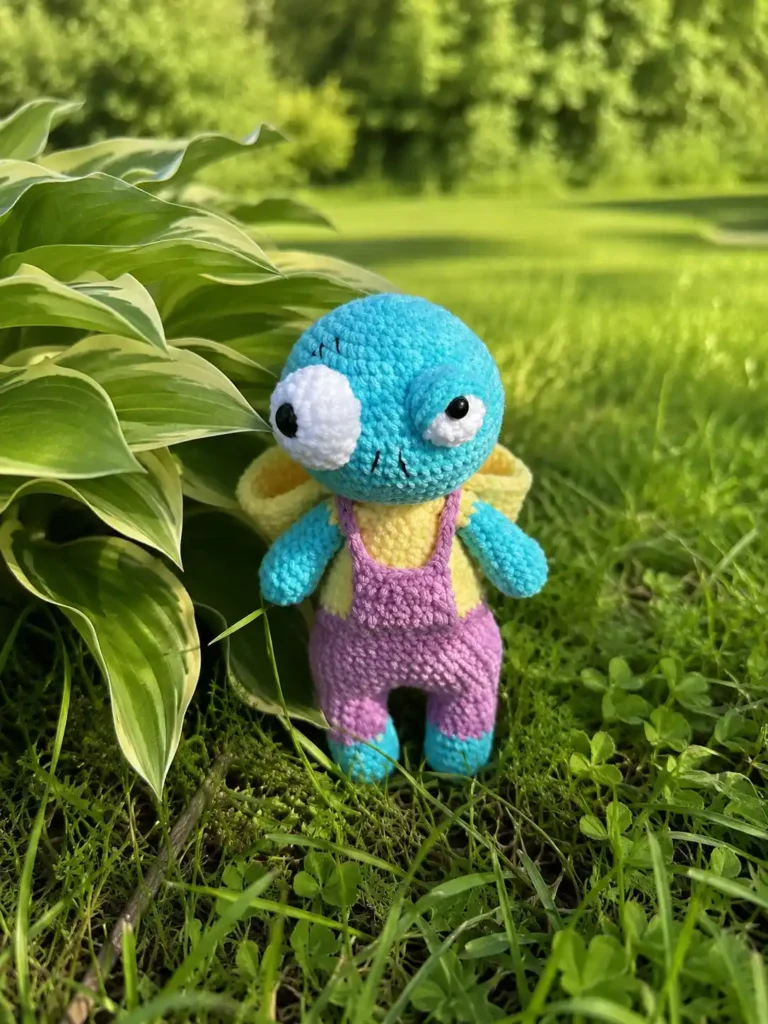 Front view of the Oswin zombie amigurumi doll standing in a grassy garden, showing his round blue-green head with his pale yellow hood pulled back onto his shoulders.