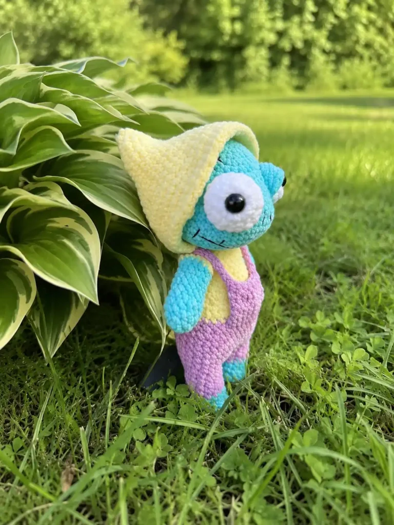 Side profile of a completed Oswin zombie amigurumi plush showing the pointed yellow hood, light blue-green skin, stitched smile, and pink overalls while leaning against garden leaves.
