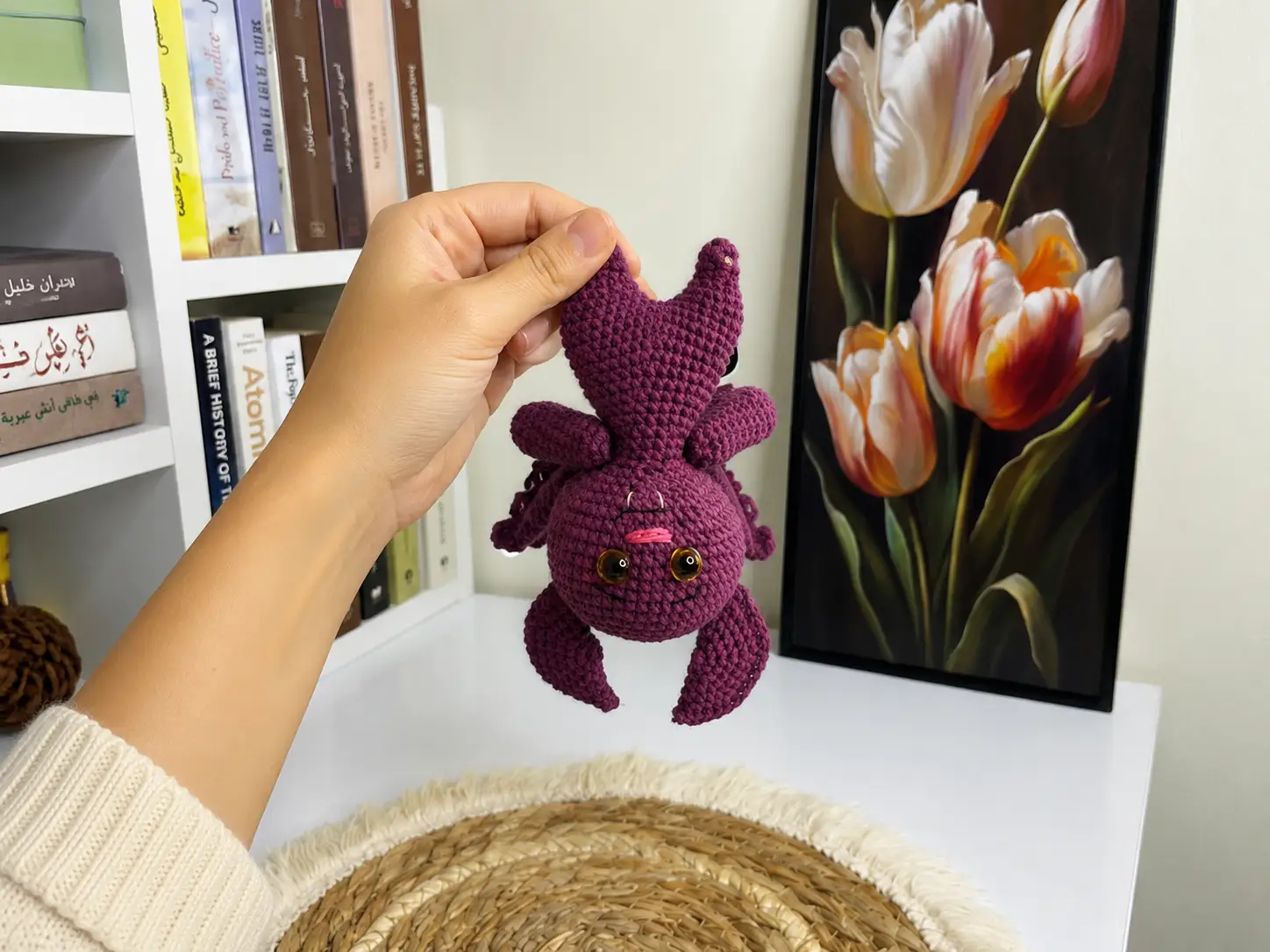 Hand holding a purple yarn halloween bat crochet pattern toy indoors near a sunlit window.