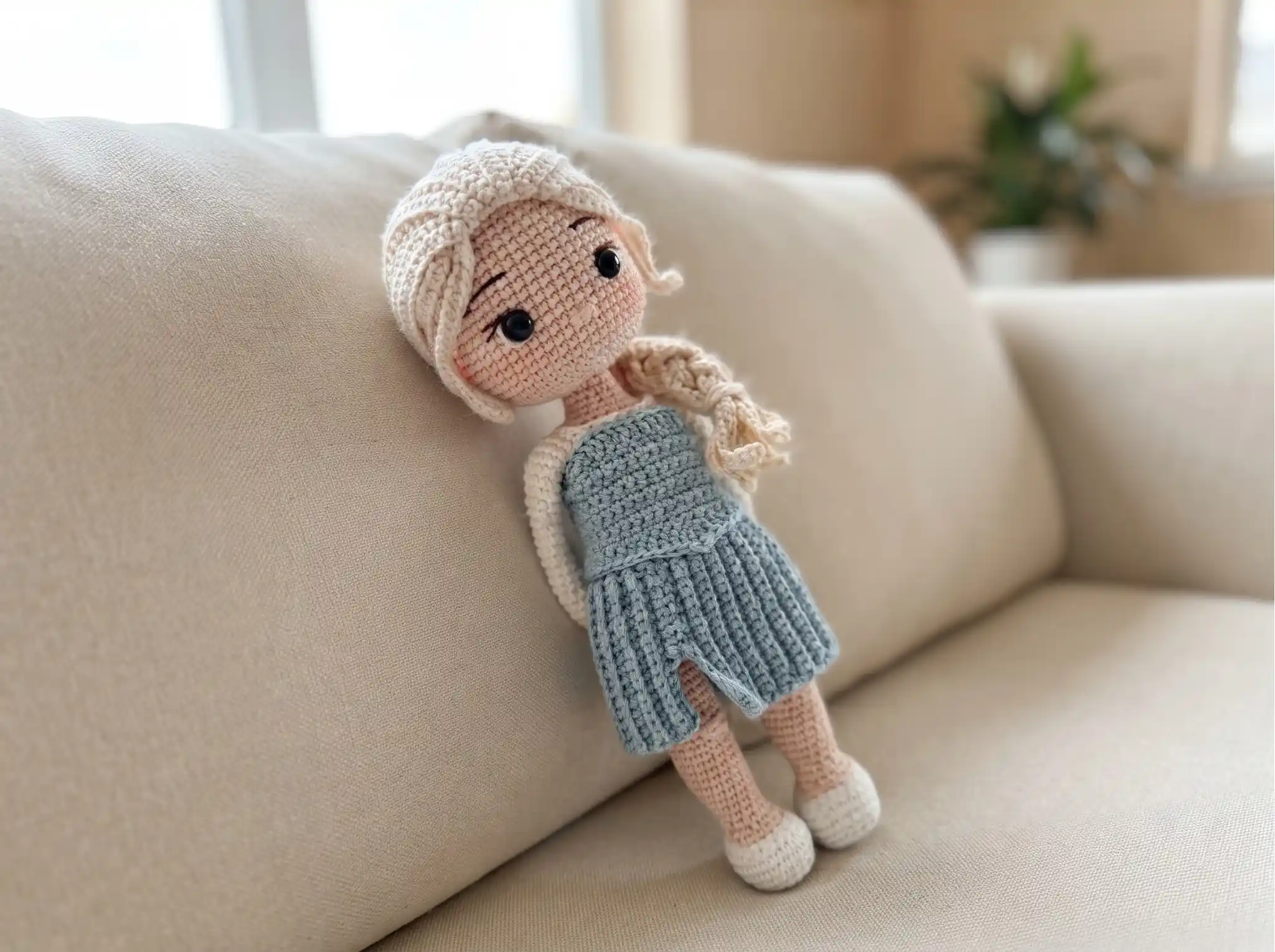 Crochet ice queen amigurumi with blonde braided hair and a blue dress resting on a beige sofa.
