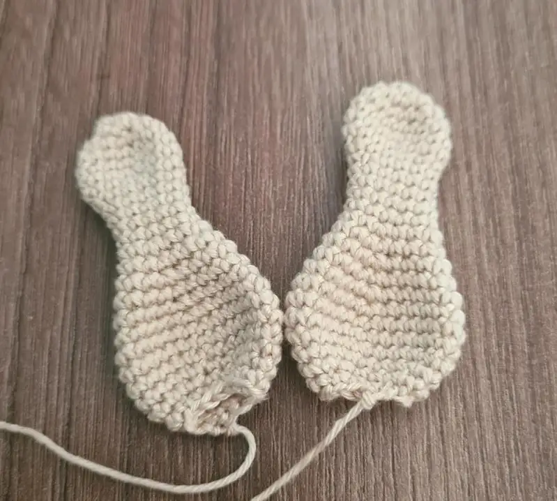 Two flat, beige crocheted amigurumi ears shaped like double spoons, resting on a wooden surface with long yarn tails left for sewing.