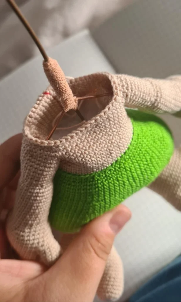 A headless crocheted amigurumi figure standing against a grid paper background. The figure has large three-toed feet, green pants, a beige torso, and long arms with jointed fingers. A wire extends vertically from the neck opening.