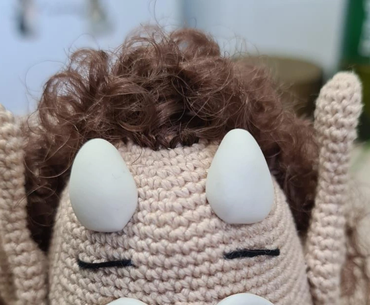 Extreme close-up of a crocheted beige amigurumi head showing attached curly brown hair, solid white sculpted eyes, and straight black embroidered eyebrows.