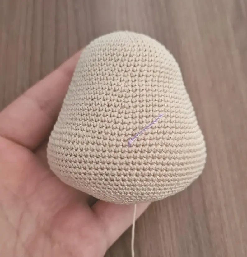 A pear-shaped, beige crocheted amigurumi head resting in a hand, with a small piece of purple yarn marking a stitch near the center.