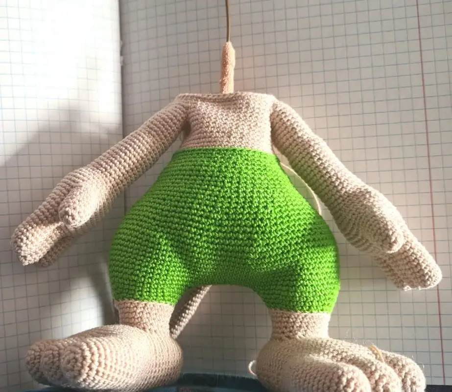 Close-up of a partially finished crocheted amigurumi body held in a hand, looking down into the open beige neck to show wires twisted together and extending upward, partially wrapped in medical tape. The lower half of the visible body features green crocheted pants.