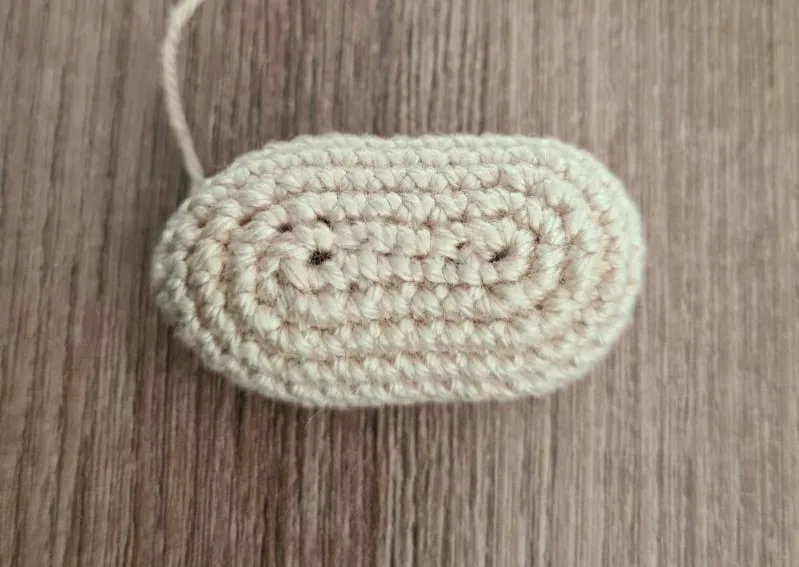 Close-up of a small, oval-shaped crocheted beige amigurumi nose resting on a wooden surface, showing continuous rounds worked around a central starting chain.