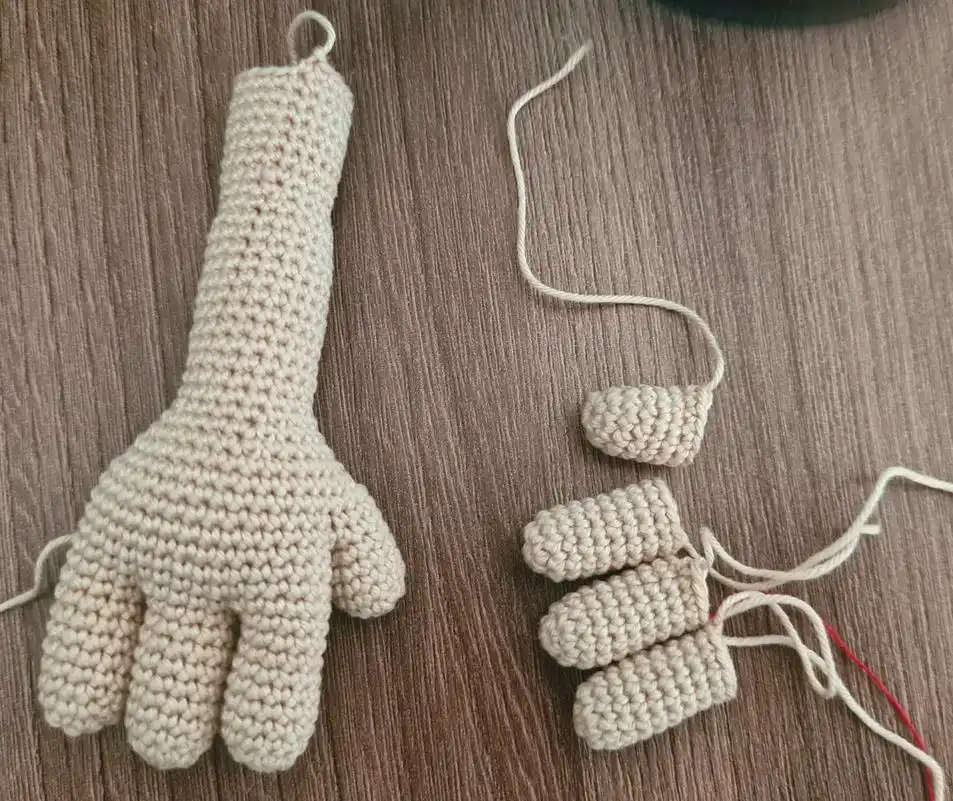 A completed crocheted beige amigurumi arm and hand with a thumb alongside three detached crocheted fingers on a wooden surface.