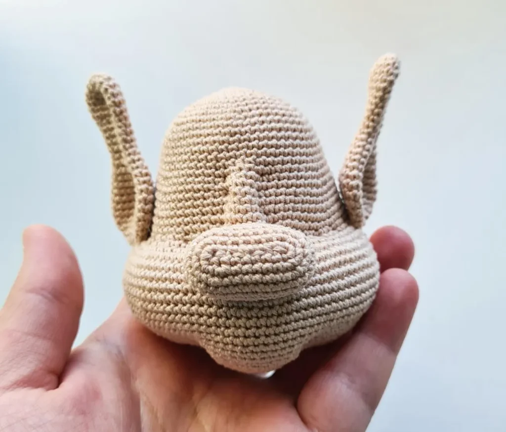 A slightly tilted, handheld photo of a crocheted beige amigurumi head featuring tall, spoon-shaped ears, a prominent attached nose, and gathered facial details against a clean, removed background.