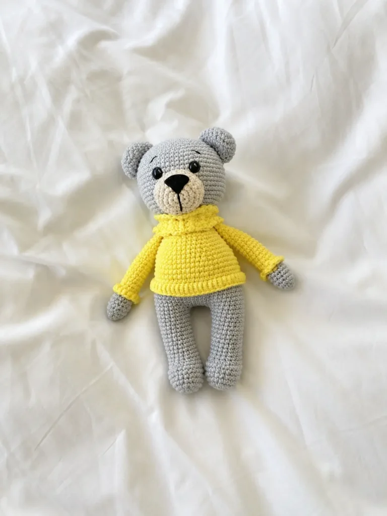 Gray crochet amigurumi bear in a bright yellow turtleneck sweater lying flat on a white duvet bedsheet