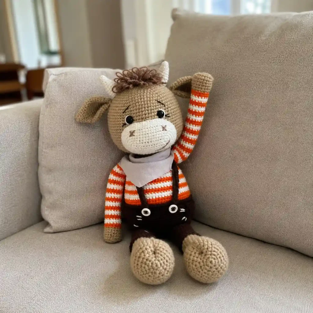 A handcrafted crochet plush bull named Mumuka sitting on a beige couch cushion with one arm raised, featuring a soft cream muzzle, dark brown overall shorts, and small white horns.