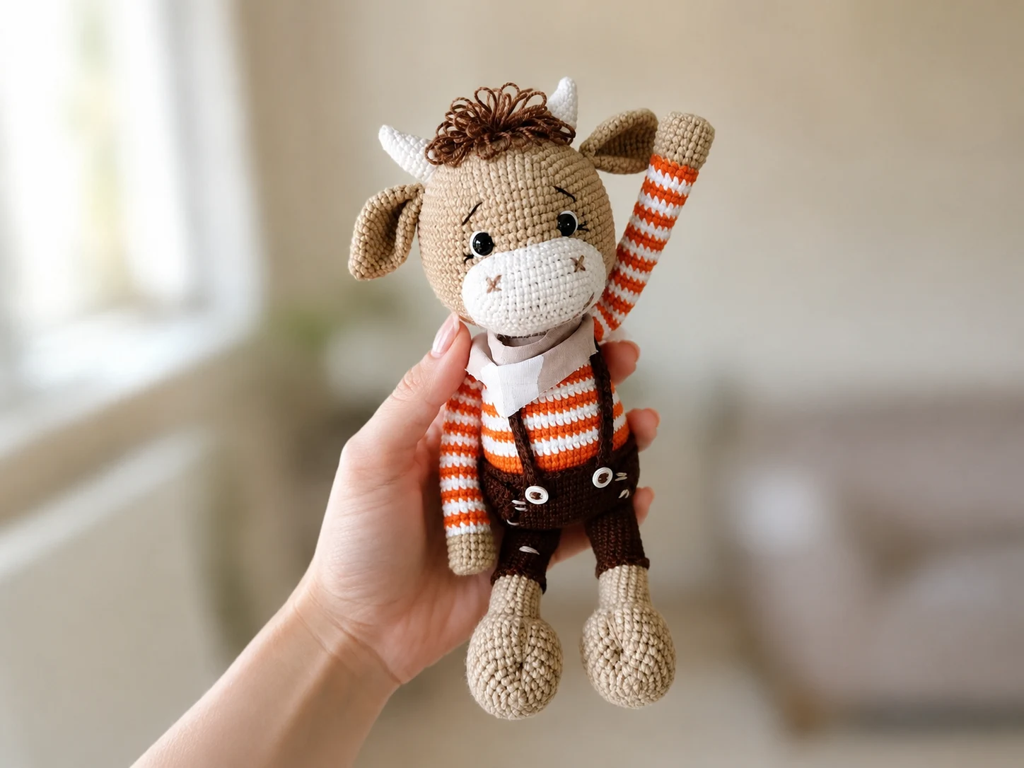 Hand holding brown crocheted bull with orange striped sweater and overalls against a soft indoor background.