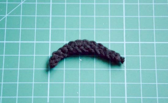 A small, curved piece of black crocheted yarn resting on a green, gridded cutting mat. The piece is thicker in the center and tapers at both ends, resembling a cartoon eyebrow.